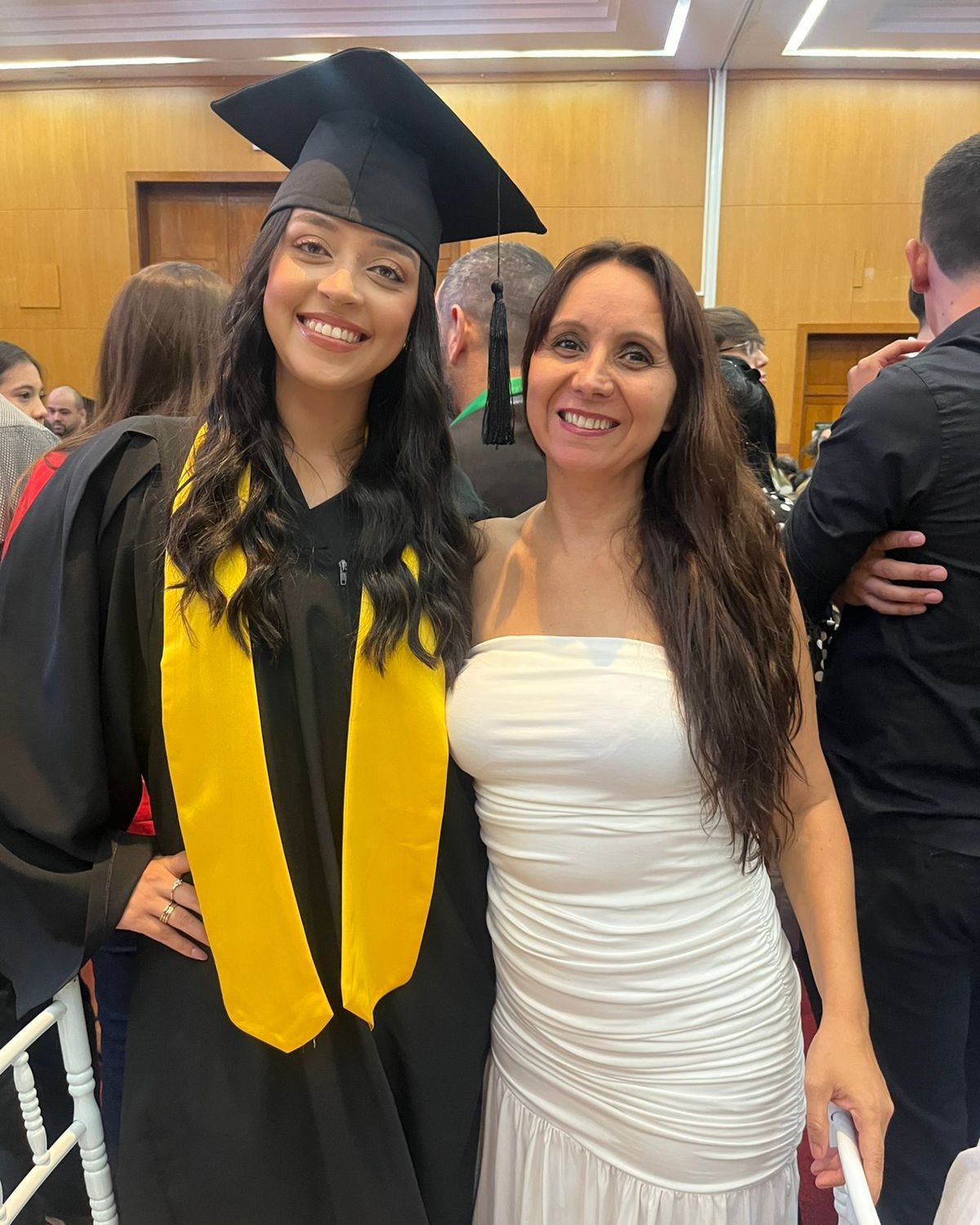 Formanda de beca e capelo sorrindo ao lado de outra mulher durante a cerimônia de formatura